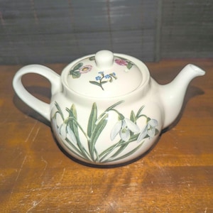 May include: A white ceramic teapot with a lid, handle, and spout. The teapot is decorated with a floral design featuring snowdrop flowers and green leaves. The lid has a small knob and additional floral accents.