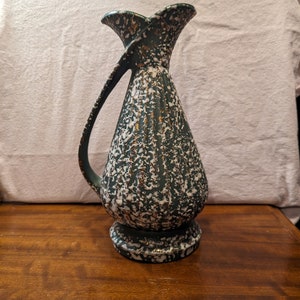 May include: A green and white ceramic vase with gold accents. The vase has a unique shape with a curved handle and a wide mouth.