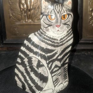 May include: A hand-painted wooden cat figurine featuring a grey and white tabby coat with bold black stripes. The cat has bright orange eyes, a small pink nose, and pointed ears, depicted in a seated position with its tail curled around its side.
