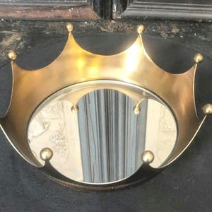 May include: A circular tray featuring a metallic gold-toned rim shaped like a crown with pointed arches and small spherical finials at each tip. The base of the tray consists of a reflective circular mirror surface that shows a clear reflection.