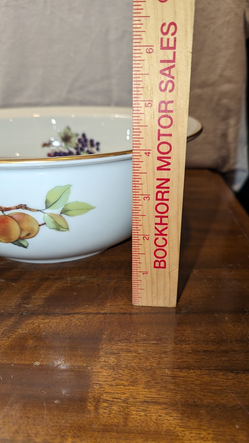 K&ouml;nnte beinhalten: Eine wei&szlig;e Porzellanschale mit goldfarbenem Rand und einem Blumendesign mit &Auml;pfeln und Bl&auml;ttern. Die Schale hat einen Durchmesser von 10 cm und steht auf einer Holzoberfl&auml;che neben einem Lineal.
