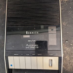May include: A black cassette tape player with the brand name "ZENITH" and the text "ac/dc Automatic Level Control-Off". The player has buttons for rewind, fast forward, play, stop, and volume.