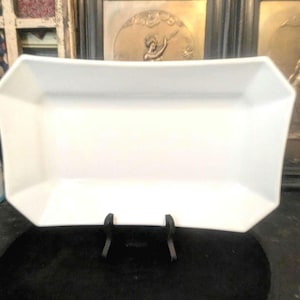 May include: A rectangular white ceramic serving dish with angled sides and a smooth finish. The dish is displayed on a black stand, featuring a wide, flat base and flared edges that create a clean, geometric silhouette for food presentation.