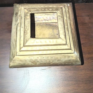 May include: A square, gold-colored metal box with a raised, decorative border. The box has a square opening in the center.