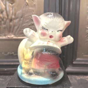 May include: A white ceramic figurine of a small, anthropomorphic creature with large ears, wearing a red and white outfit and holding a yellow bunny. The figurine is standing on a blue base and is displayed on a black pedestal.