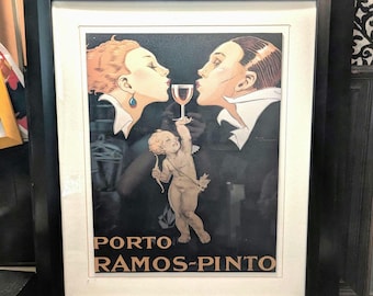 Op de afbeelding: Ingelijste vintage advertentie voor Porto Ramos-Pinto. Het kunstwerk toont twee figuren die kussen boven een wijnglas, met een cherubijn die een pijl en boog vasthoudt. De tekst "PORTO RAMOS-PINTO" wordt weergegeven in gouden letters.