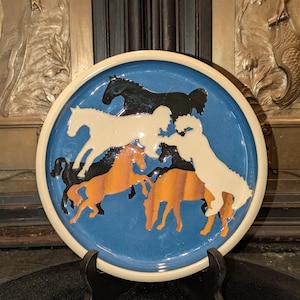 May include: A ceramic plate with a blue center and a cream-colored rim. The plate features a design of six horses in shades of black, white, and orange. The horses are arranged in a circular pattern, creating a dynamic and artistic composition. The plate is displayed on a black stand.