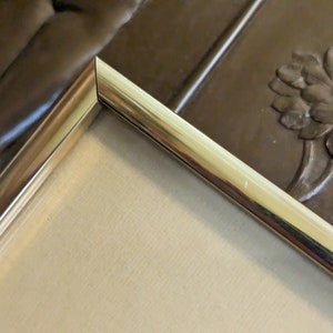 May include: Close-up of the corner of a gold-coloured picture frame. The frame has a smooth, polished finish and a slightly raised edge.
