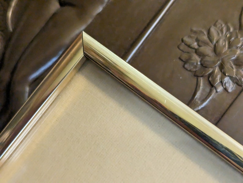 May include: Close-up of the corner of a gold-coloured picture frame. The frame has a smooth, polished finish and a slightly raised edge.
