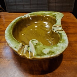 May include: A green and brown ceramic ashtray with a unique, wavy design. The ashtray has a wide, shallow bowl and a raised rim.