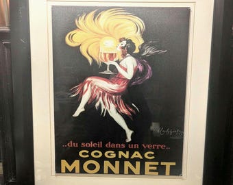 May include: Framed vintage Cognac Monnet advertisement. The artwork features a woman in a red dress holding a glass of cognac, with a large yellow sun-like halo. The text reads "...du soleil dans un verre.. COGNAC MONNET."