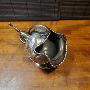 May include: A silver-plated creamer or gravy boat with a decorative floral design. The boat has a spout and a handle.