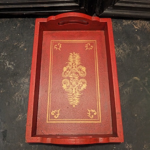 May include: A red wooden serving tray with a gold painted design. The tray has a raised edge and a handle on the top.