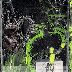 Peut inclure: Une œuvre d'art encadrée avec un fond sombre et texturé et des tourbillons de peinture vert néon. Une sculpture d'insecte détaillée et de couleur foncée est montée à l'intérieur du cadre. L'œuvre comprend une étiquette argentée avec le texte "TZOLKINSTUDIO ART".