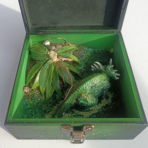 May include: A green wooden box with a hinged lid, open to reveal a miniature dinosaur figurine and a small green plant. The dinosaur is green and has a spiky back. The plant is a small, leafy plant with a brown stem.