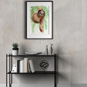 May include: A framed watercolour painting of a brown sloth hanging from a branch with green leaves. The sloth has a friendly expression and is looking directly at the viewer.