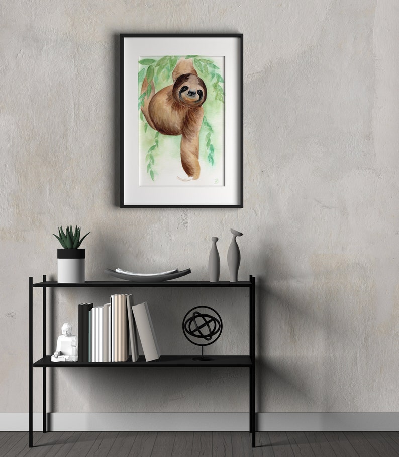 May include: A framed watercolour painting of a brown sloth hanging from a branch with green leaves. The sloth has a friendly expression and is looking directly at the viewer.