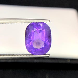 Puede incluir: Una gema p&uacute;rpura ovalada y facetada, sujeta con pinzas. La superficie de la gema refleja la luz, mostrando su estructura interna y variaciones de color. El color de la gema var&iacute;a de un violeta intenso a un tono lavanda m&aacute;s claro.