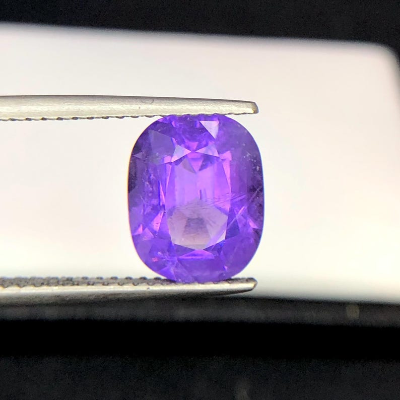 Puede incluir: Una gema p&uacute;rpura ovalada y facetada, sujeta con pinzas. La superficie de la gema refleja la luz, mostrando su estructura interna y variaciones de color. El color de la gema var&iacute;a de un violeta intenso a un tono lavanda m&aacute;s claro.