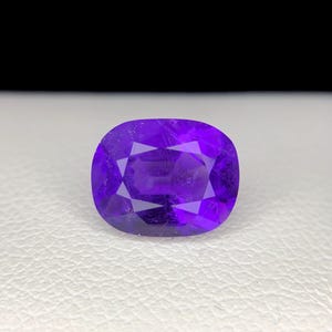 Puede incluir: Una gema facetada de color p&uacute;rpura intenso, de forma ovalada. La gema tiene una superficie pulida con m&uacute;ltiples facetas que reflejan la luz. La piedra se encuentra sobre una superficie blanca con un fondo negro.