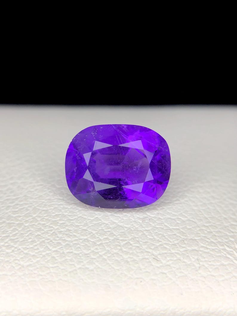 Puede incluir: Una gema facetada de color p&uacute;rpura intenso, de forma ovalada. La gema tiene una superficie pulida con m&uacute;ltiples facetas que reflejan la luz. La piedra se encuentra sobre una superficie blanca con un fondo negro.