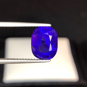 Puede incluir: Una gema azul oscuro, facetada y de forma ovalada, es sostenida por unas pinzas plateadas. El tallado brillante de la gema refleja la luz, mostrando su rico color. El fondo oscuro resalta el vibrante tono de la gema.