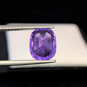 Puede incluir: Una gema facetada, de forma ovalada, de color p&uacute;rpura intenso, sujeta con pinzas. La gema tiene una superficie pulida con estructuras internas visibles y una calidad brillante y reflectante. El fondo es un suave degradado de blanco y negro.