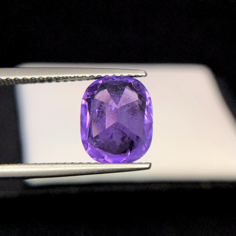 Puede incluir: Una gema facetada, de forma ovalada, de color p&uacute;rpura intenso, sujeta con pinzas. La gema tiene una superficie pulida con estructuras internas visibles y una calidad brillante y reflectante. El fondo es un suave degradado de blanco y negro.