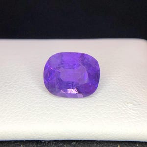 Puede incluir: Una gema p&uacute;rpura facetada, de forma ovalada. La gema es de un tono violeta vibrante, con estructuras internas visibles y una superficie pulida. La piedra est&aacute; sobre un fondo blanco, con un fondo oscuro.