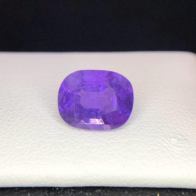 Puede incluir: Una gema p&uacute;rpura facetada, de forma ovalada. La gema es de un tono violeta vibrante, con estructuras internas visibles y una superficie pulida. La piedra est&aacute; sobre un fondo blanco, con un fondo oscuro.