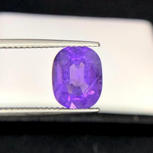 Puede incluir: Una gema p&uacute;rpura facetada, de forma ovalada, es sostenida por unas pinzas. La gema tiene un corte brillante, que refleja la luz y muestra su color violeta intenso. El fondo es blanco suave, resaltando la claridad y el brillo.