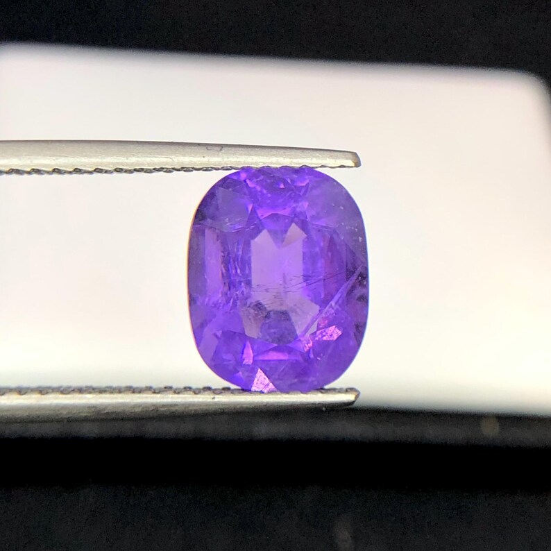 Puede incluir: Una gema p&uacute;rpura facetada, de forma ovalada, es sostenida por unas pinzas. La gema tiene un corte brillante, que refleja la luz y muestra su color violeta intenso. El fondo es blanco suave, resaltando la claridad y el brillo.