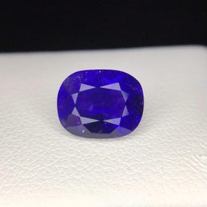 Puede incluir: Una gema azul oscuro facetada, de forma ovalada. La superficie pulida tiene m&uacute;ltiples facetas que reflejan la luz, creando un efecto brillante. La piedra est&aacute; sobre un fondo blanco.