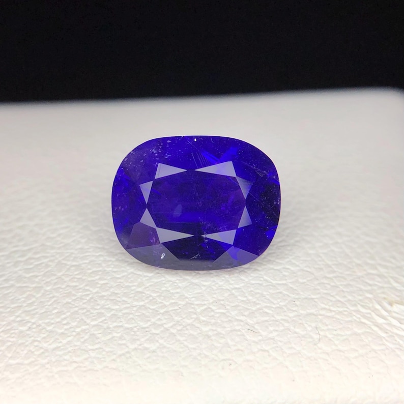 Puede incluir: Una gema azul oscuro facetada, de forma ovalada. La superficie pulida tiene m&uacute;ltiples facetas que reflejan la luz, creando un efecto brillante. La piedra est&aacute; sobre un fondo blanco.