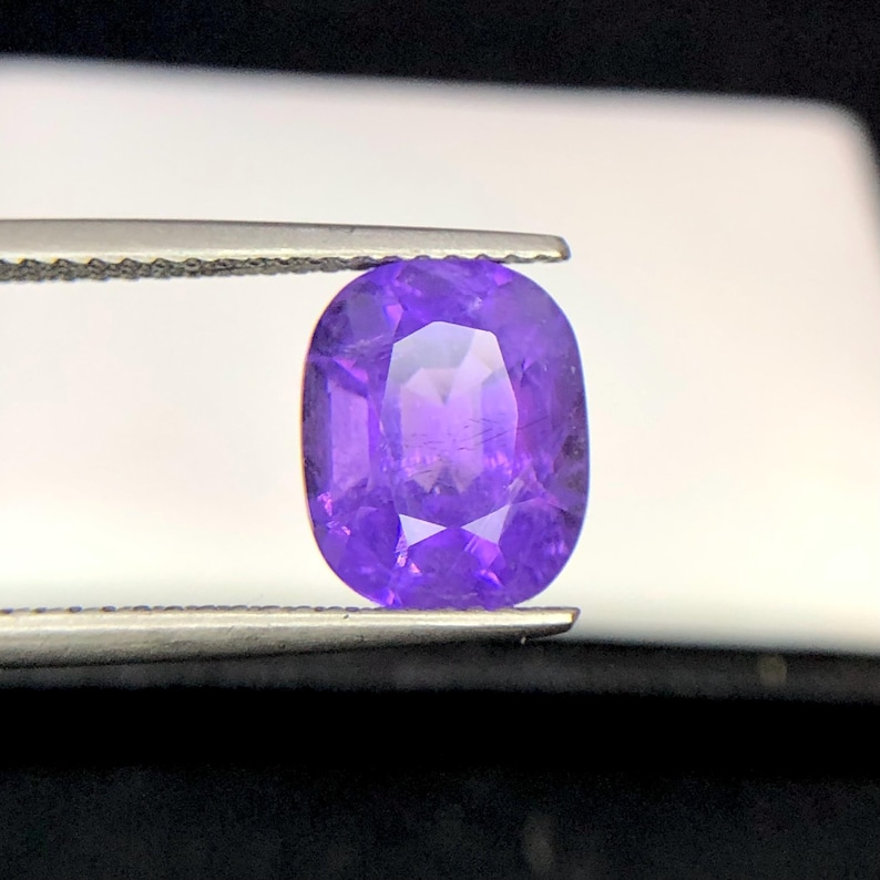 Puede incluir: Una gema p&uacute;rpura de corte ovalado, sujeta con pinzas. La gema tiene una superficie pulida con m&uacute;ltiples facetas, que reflejan la luz. El color es un violeta intenso, con un ligero degradado. La piedra preciosa es el punto focal, sobre un fondo borroso.