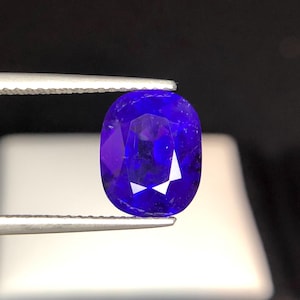 Puede incluir: Una gema facetada, de forma ovalada, de color azul intenso, sujeta con pinzas. La gema tiene una superficie pulida con facetas internas que reflejan la luz, creando un brillo. El fondo oscuro resalta el color de la gema.