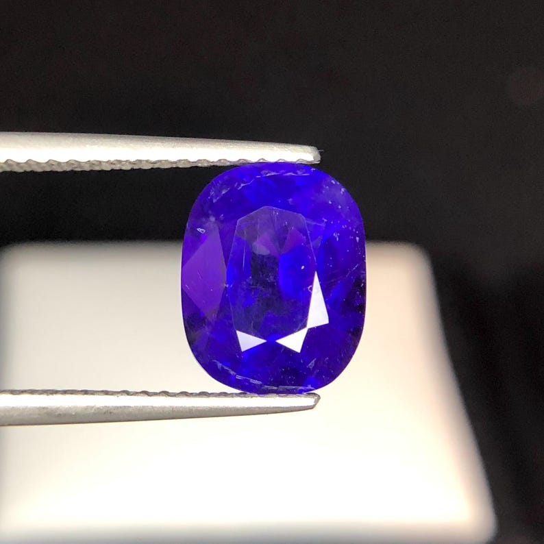 Puede incluir: Una gema facetada, de forma ovalada, de color azul intenso, sujeta con pinzas. La gema tiene una superficie pulida con facetas internas que reflejan la luz, creando un brillo. El fondo oscuro resalta el color de la gema.