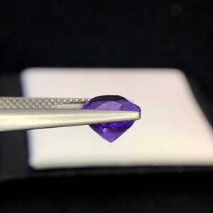 Puede incluir: Una gema facetada de color p&uacute;rpura oscuro sostenida por unas pinzas plateadas. La gema tiene un corte brillante, que refleja la luz. El fondo es una mezcla de blanco y negro, resaltando el color y la claridad de la joya. La gema es de color violeta oscuro.