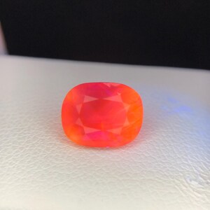 Puede incluir: Una gema vibrante, de talla coj&iacute;n, con una mezcla ardiente de tonos rojos y naranjas. La gema se encuentra sobre una superficie blanca, mostrando sus facetas y brillo interno. La forma de la gema es un rect&aacute;ngulo redondeado.