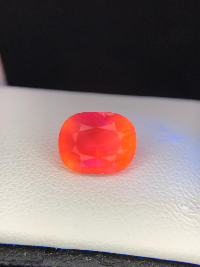Puede incluir: Una gema vibrante, de talla coj&iacute;n, con una mezcla ardiente de tonos rojos y naranjas. La gema se encuentra sobre una superficie blanca, mostrando sus facetas y brillo interno. La forma de la gema es un rect&aacute;ngulo redondeado.