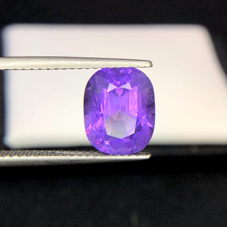 Puede incluir: Una gema p&uacute;rpura de talla ovalada y facetada, sujeta con pinzas. La superficie de la gema refleja la luz, mostrando su estructura interna y variaciones de color. La gema es de un tono violeta intenso.