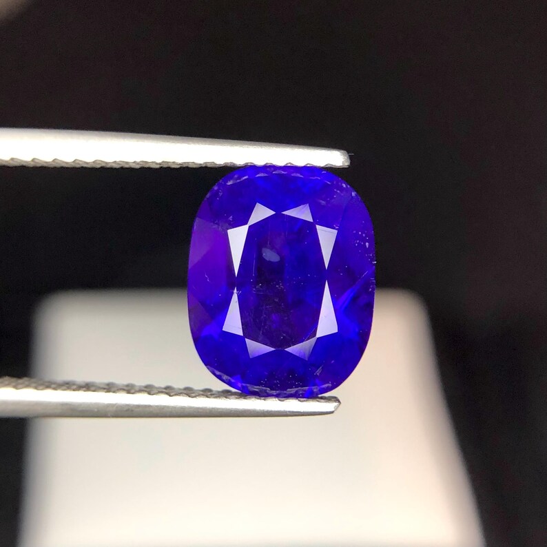 Puede incluir: Una gema ovalada de color azul intenso es sostenida por unas pinzas. La gema presenta un complejo patr&oacute;n de facetas internas, creando un efecto de estallido estelar. El fondo es oscuro, resaltando el brillo de la gema.