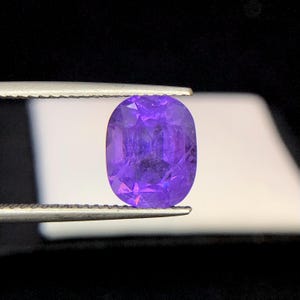 Puede incluir: Una gema p&uacute;rpura facetada, de forma ovalada, es sostenida por unas pinzas sobre un fondo blanco y negro. La gema tiene un corte brillante, mostrando su estructura interna y color. La gema es de un tono violeta vibrante.
