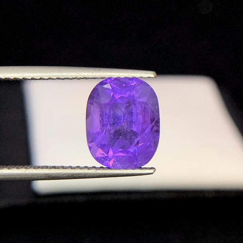 Puede incluir: Una gema p&uacute;rpura facetada, de forma ovalada, es sostenida por unas pinzas sobre un fondo blanco y negro. La gema tiene un corte brillante, mostrando su estructura interna y color. La gema es de un tono violeta vibrante.