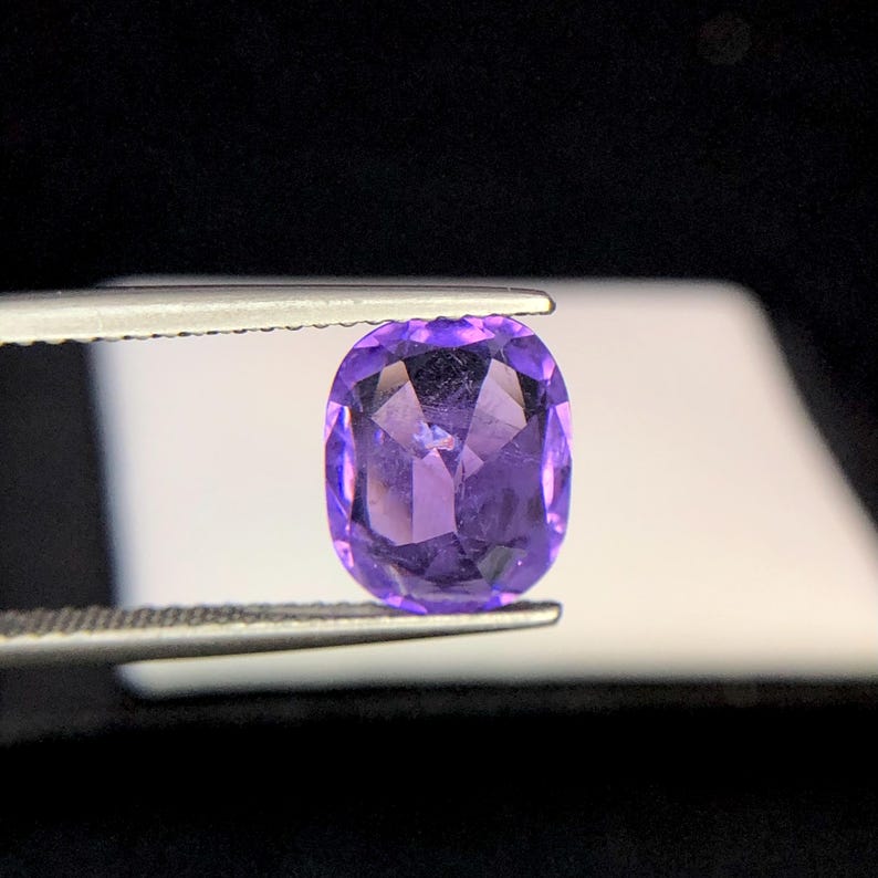 Puede incluir: Una gema p&uacute;rpura facetada, de corte ovalado, sujeta con pinzas. La gema tiene un tono violeta intenso y una superficie brillante y reflectante. El fondo es un degradado de negro y blanco, resaltando el color y la claridad de la gema.