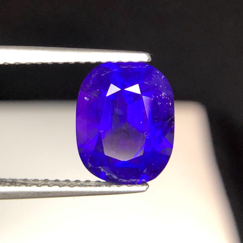 Puede incluir: Una gema azul oscuro, tallada en forma ovalada, sujeta con pinzas. La gema tiene m&uacute;ltiples facetas que reflejan la luz. El color es un azul rico y saturado con un ligero matiz p&uacute;rpura. La piedra preciosa se muestra sobre un fondo oscuro.