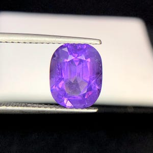 Puede incluir: Una gema p&uacute;rpura facetada de corte ovalado, sujeta con pinzas. La gema tiene una superficie brillante y reflectante con m&uacute;ltiples facetas que captan la luz. El color es un p&uacute;rpura profundo y rico, con tonos m&aacute;s claros visibles.