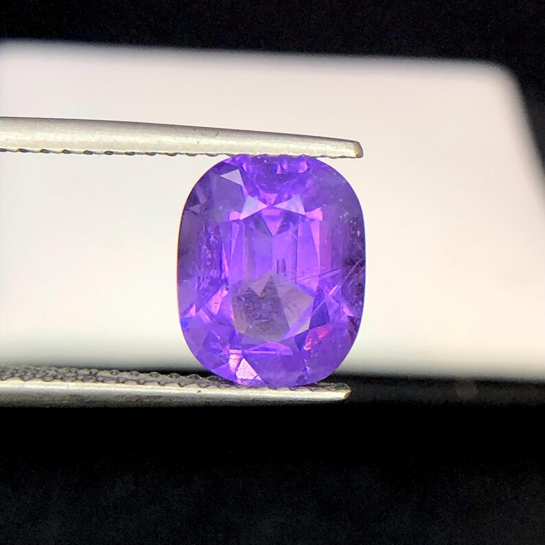 Puede incluir: Una gema p&uacute;rpura facetada de corte ovalado, sujeta con pinzas. La gema tiene una superficie brillante y reflectante con m&uacute;ltiples facetas que captan la luz. El color es un p&uacute;rpura profundo y rico, con tonos m&aacute;s claros visibles.