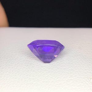 Puede incluir: Una gema facetada de color p&uacute;rpura intenso con forma rectangular. La gema tiene m&uacute;ltiples facetas que reflejan la luz, creando un efecto brillante. La piedra est&aacute; colocada sobre una superficie blanca, con un fondo negro.