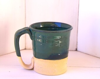 May include: A green and cream ceramic mug with a handle. The mug is glazed in a dark green color with a cream colored band around the bottom.